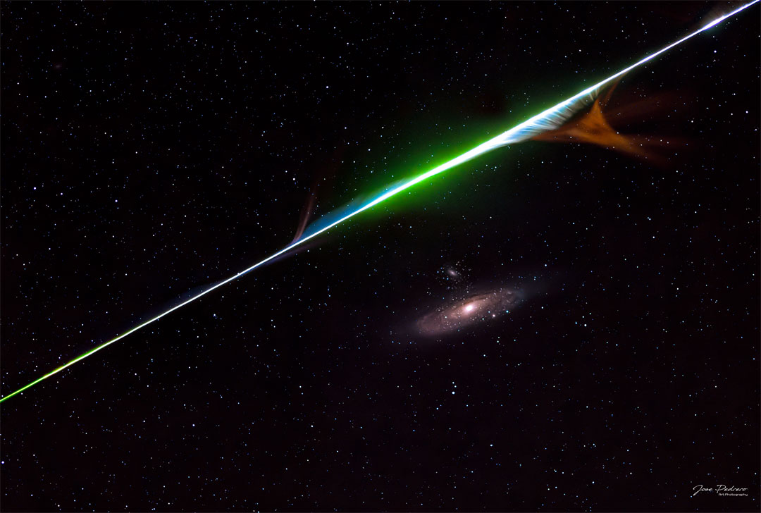 Ein Meteor blitzt diagonal durchs Bild und verfehlt knapp die Andromedagalaxie. Die Meteorspur ist farbig und zieht Schlieren.