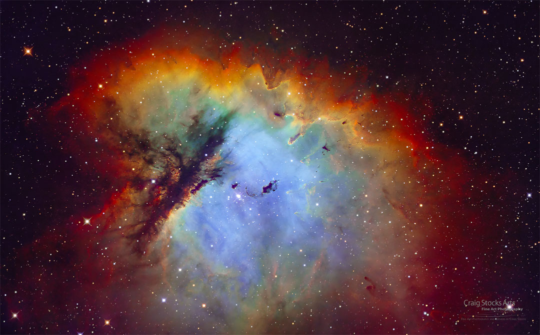 Der Emissionsnebel NGC 281 im Bild leuchtet innen blau und ist von einem orange-roten Grat umgeben. Links ragt eine dunkle Wolke in den Nebel hinein. Der Nebel erinnert an Pacman.