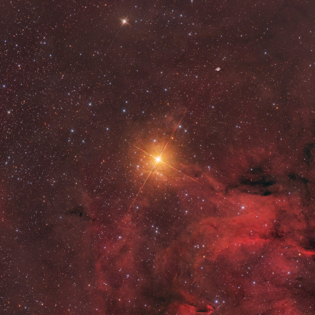 In der Mitte leuchtet ein orange-rötlicher Stern, er ist der hellste Stern im Bild. Nach rechts unten breiten sich rote Nebelwolken aus.