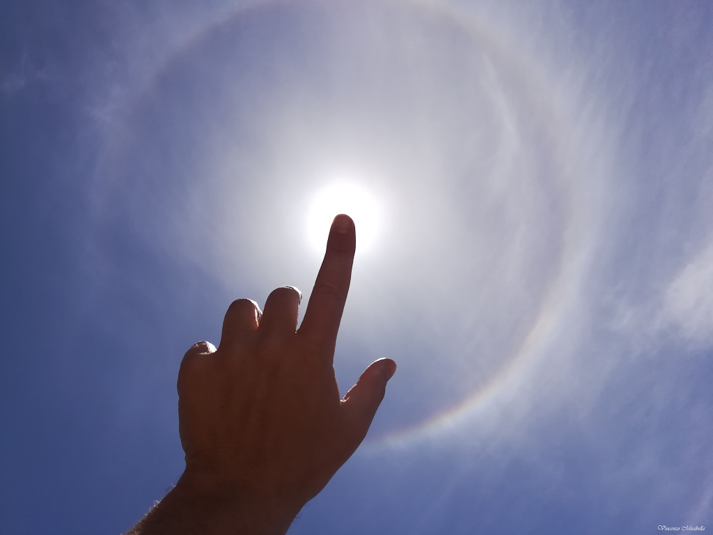 Eine Hand hebt den Zeigefinger zur Bildmitte, hinter der Fingerspitze ist die Sonne. Um die Sonne verläuft ein 22-Grad-Halo.