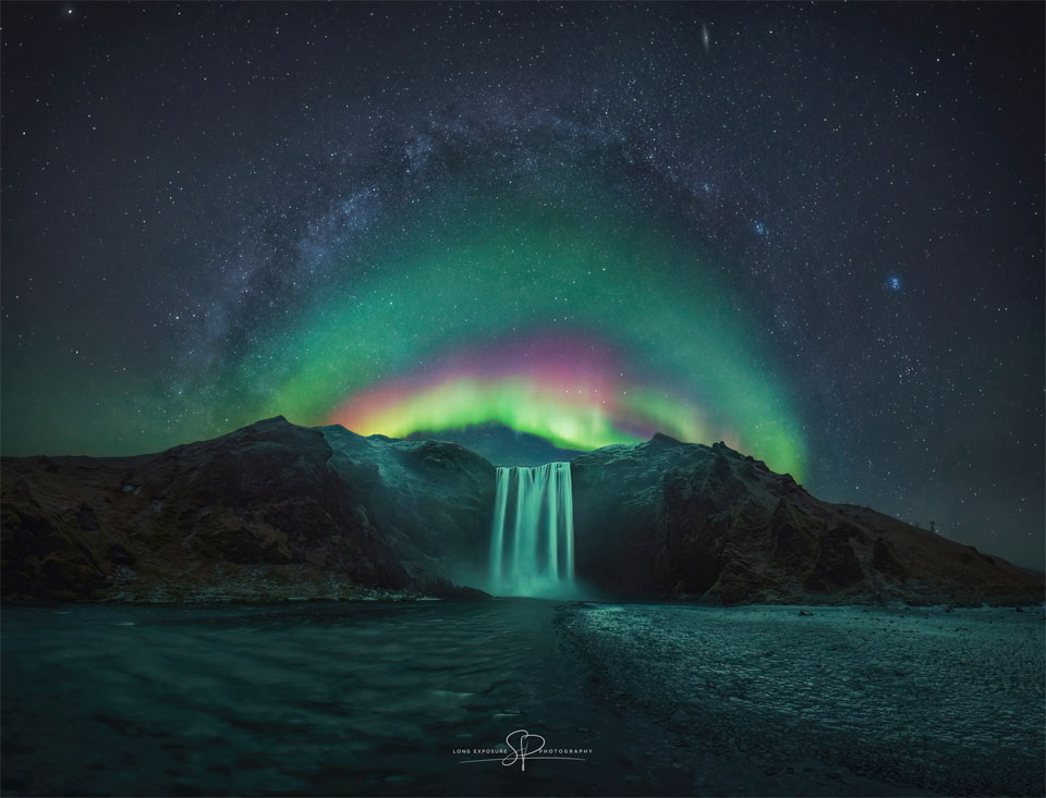 In der Bildmitte ist ein Wasserfall unter einem Sternenhimmel zu sehen. Über dem Wasserfall wölbt sich ein buntes Polarlicht. Über dem Polarlicht wölbt sich das zentrale Band der Milchstraße.