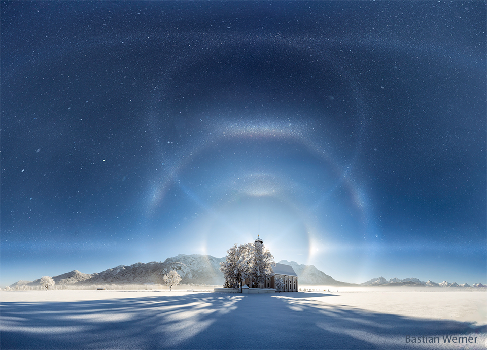 In der Ferne steht ein Gebäude auf weißem Schnee mit Bergen im Hintergrund. Darüber ist ein Himmel voller Eiskristalle. Der Nachthimmel wird von zahlreichen geschwungenen Eiskristallen überlagert, die das Mondlicht im Hintergrund reflektieren.