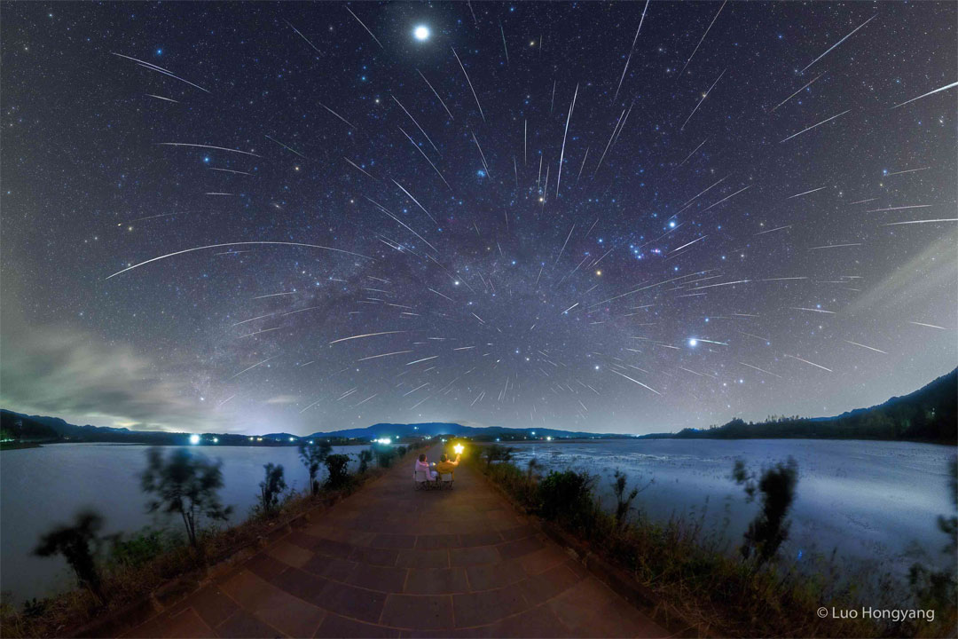 Über einem See strömen zahllose Meteore der Geminiden vom Himmel. Im Hintergrund ist das Sternbild Orion und die Milchstraße zu erkennen.