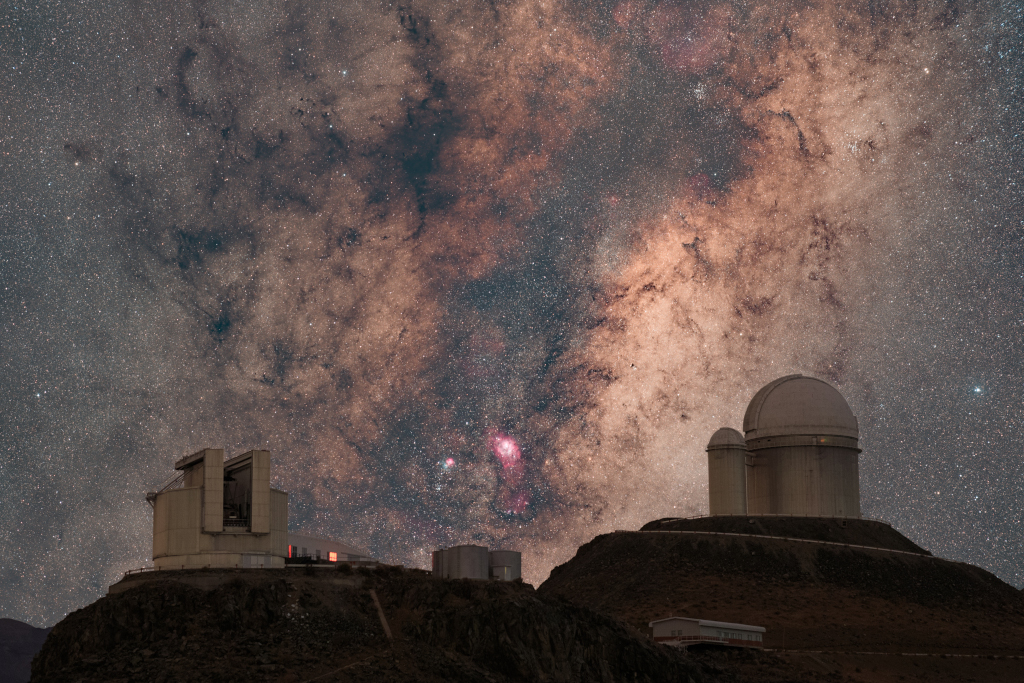 Hinter mehreren Teleskopkuppeln auf einem Berg breitet sich die Milchstraße mit Dunkelwolken aus.