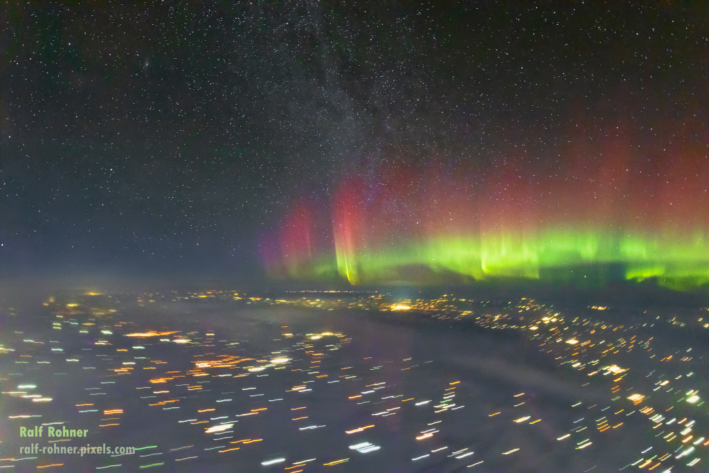 Rechts im Bild über dem Horizont leuchten Polarlichter, unten grün und darüber dunkelrot. Über dem Horizont sind zart gesprenkelte Sterne, unten auf der Erde sind zahlreiche Lichtspuren.