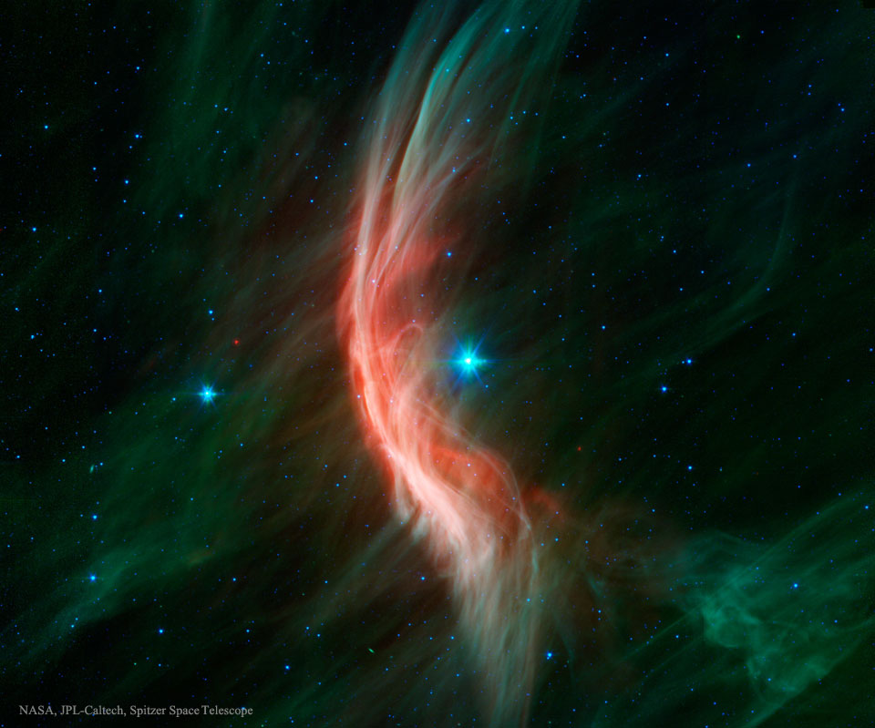Links neben dem Stern in der Mitte leuchtet ein roter Nebelschleier mit grünen Enden, der wie eine Bugwelle um den Stern gekrümmt ist.