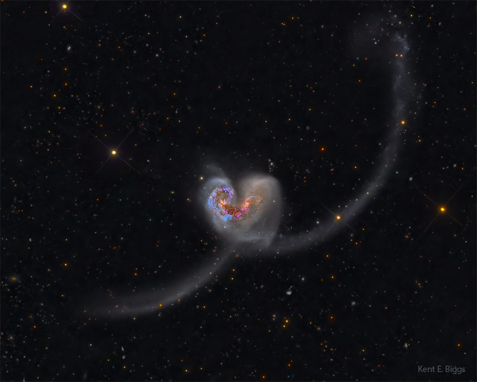 Zwei Galaxien kollidieren und bilden dabei eine Herzform. Nach links unten und rects oben verlaufen geschwungene Bögen aus Sternen.
