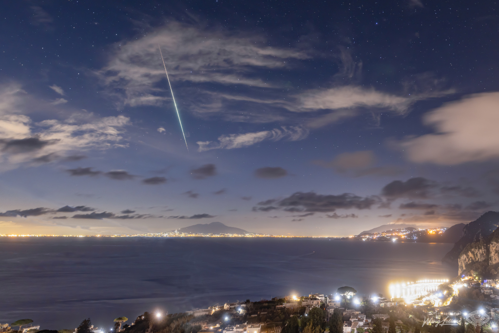 Rund um eine Bucht strahlen viele Lichter, hinter dem Gewässer ragt ein Berg über die Stadtbeleuchtung. Der Himmel ist blau mit beleuchteten Wolken, oben ist die helle Spur eines Meteors.