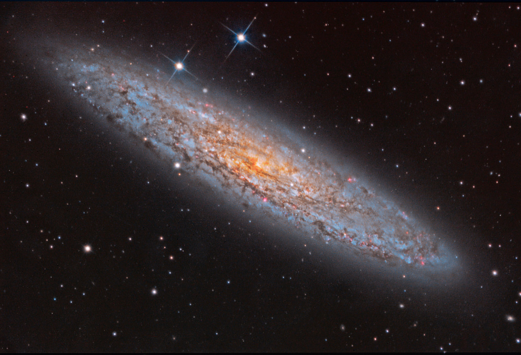 Eine sehr flache Galaxienscheibe mit vielen Dunkelwolken und Sternbildungsregionen ist schräg von oben zu sehen, sie liegt diagonal im Bild.