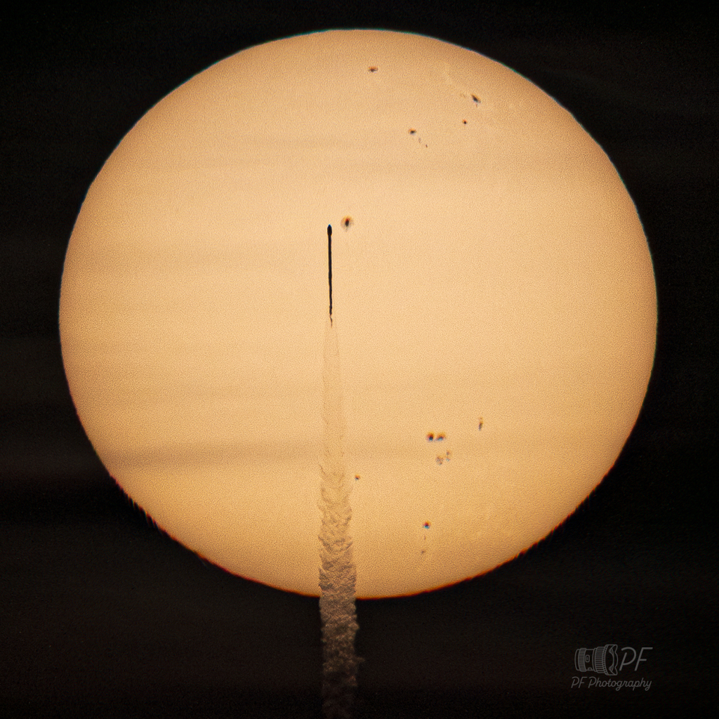 Eine Rakete zieht wie ein dunkles Streichholz über die Sonne. Auf der Sonnenoberfläche sind viele dunkle Sonnenflecken und aktive Regionen.