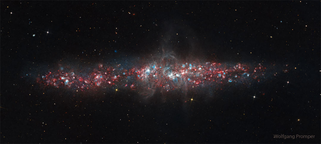 Die Galaxie NGC 55 im Bild wirkt flach. Sie schimmert wie ein Schatzkästchen voller Juwelen. Die leuchtenden Flecken sind Nebel aus Wasserstoff und Sauerstoff.