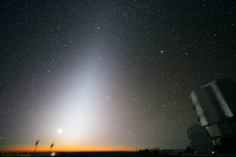 Am Horizont ragt eine Lichtpyramide hoch, dahinter ist der Himmel von relativ vielen Sternen bedeckt. Rechts steht eine Einheit des VLT und die kleine Kuppel eines Hilfsteleskops.