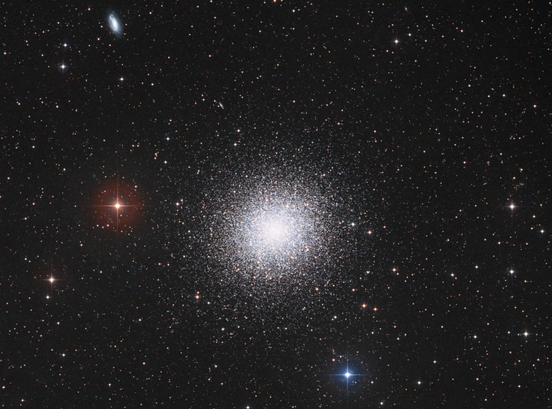 Mitten im Bild leuchtet ein großer, sehr kompakter Kugelsternhaufen. Am linken oberen Bildrand ist eine kleine Galaxie zu sehen. Im Hintergrund sind Sterne lose verteilt.
