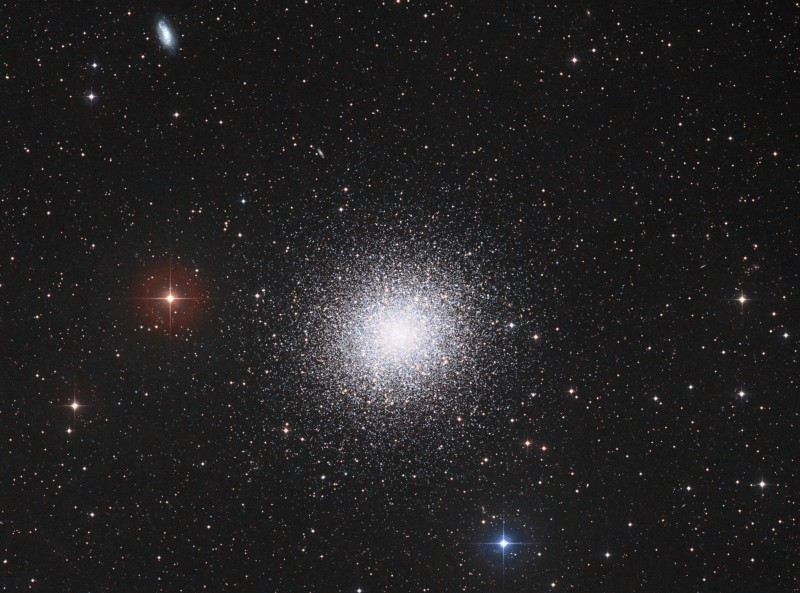 Mitten im Bild leuchtet ein großer, sehr kompakter Kugelsternhaufen. Am linken oberen Bildrand ist eine kleine Galaxie zu sehen. Im Hintergrund sind Sterne lose verteilt.