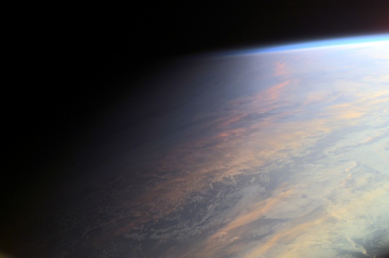 Die Erde ist von Wolken bedeckt, das Bild zeigt einen Ausschnitt mit dem Horizont oben. Der Erdrand geht mit einem blauen Streifen in den Weltraum über. In der Mitte verläuft der Schattenverlauf, links ist es schon dunkel, rechts scheint noch die Sonne.