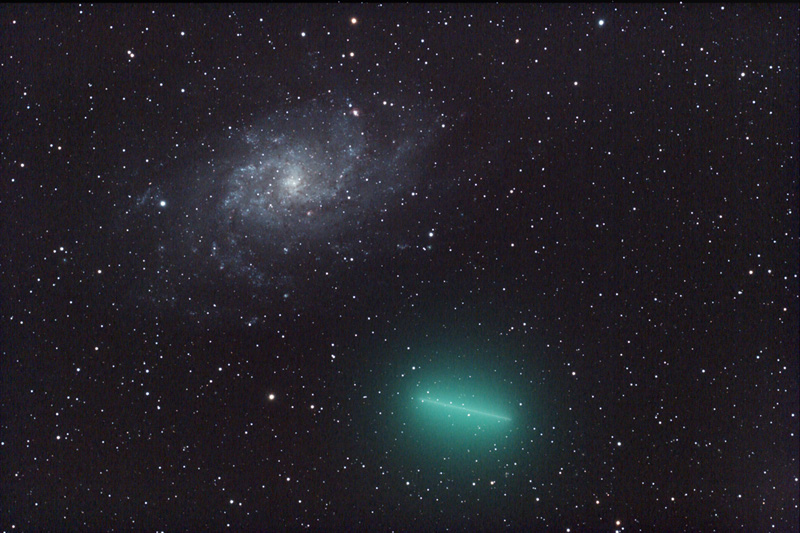 Am sternklaren Himmel begegnen sich eine Spiralgalaxie links oben und ein giftgrün leuchtender Komet unten mit türkiser Koma, der vor den Sternen eine Strichspur zieht.