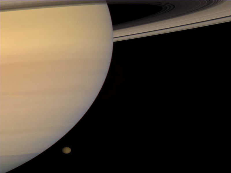 Links füllt Saturn ein Drittel des Bildes aus, am oberen Rand ist ein Teil seiner Ringe zu sehen, rechts unter dem Planeten ist im Hintergrund der Mond Titan zu sehen.