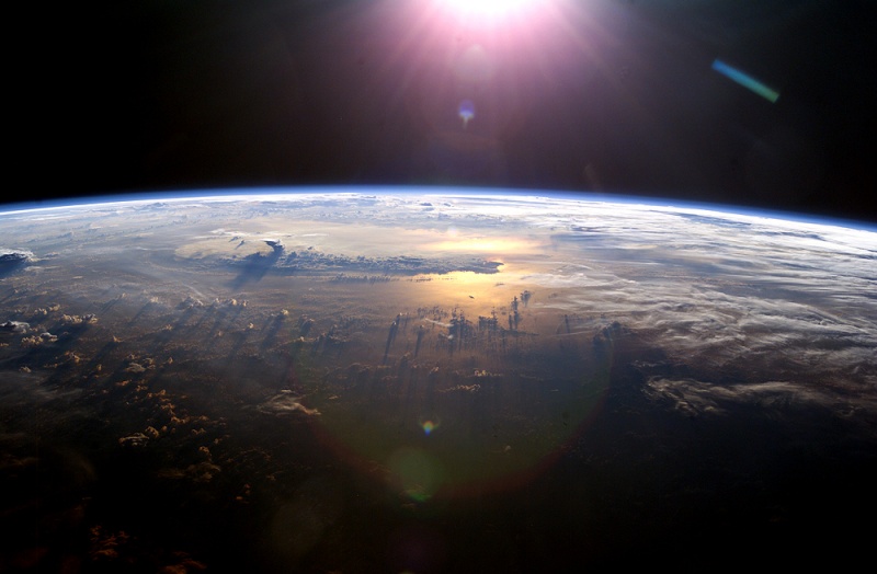 Die Erde ist aus dem Weltraum zu sehen. Im Ozean spiegelt sich die Sonne, darüber sind Wolken, am oberen Bildrand leuchtet die Sonne.