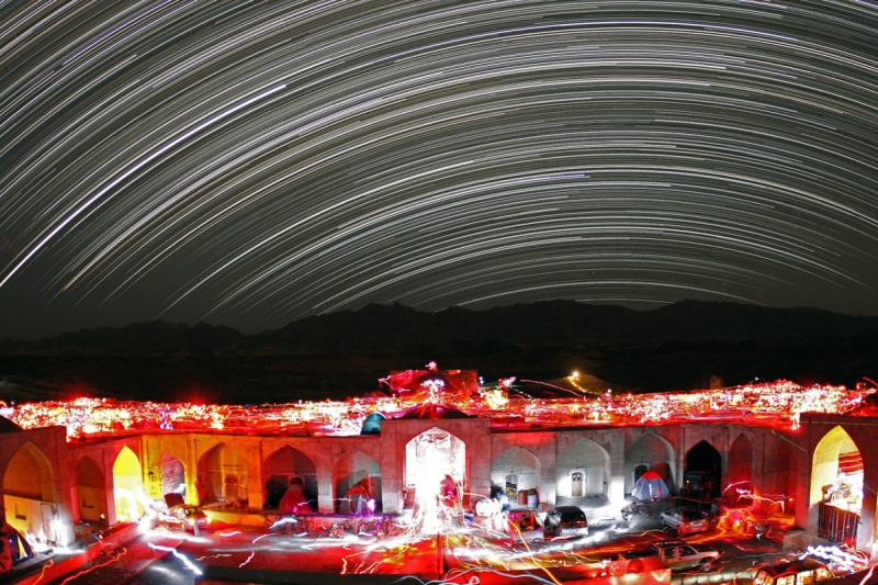 Das Bild zeigt eine Langzeitbelichtung eines Messier-Marathons. Im Vordergrund leuchten die roten und weißen Lampen der Beobachtenden in einem historischen Palast im Iran, am Himmel verlaufen die Strichspuren der Sterne.