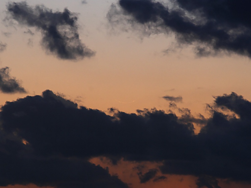 Über einer dunklen Wolkenbank ist am orangefarbenen Abendhimmel eine sehr schmale Mondsichel zu erkennen, wenn man genau hinsieht.