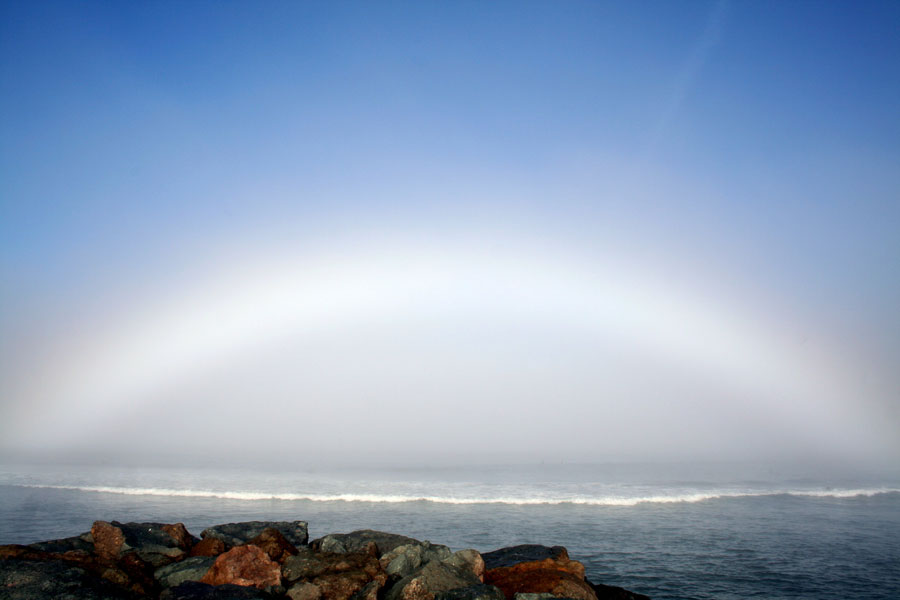 Das Bild zeigt einen weißen Nebelbogen am blauen Himmel über einem Strand.