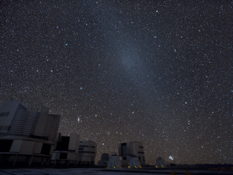 Links unten sind die Teleskope des VLT auf dem Paranal als dunkle Silhouetten vor dem fast gleich dunklen Sternenhimmel zu sehen, von links unten steigt der Gegenschein nach rechts oben steil auf.