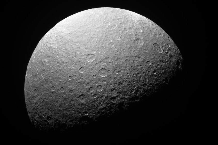 Der Mond Rhea im Bild ist von vielen Kratern übersät, die obere Hälfte ist beleuchtet, die untere ist dunkel.