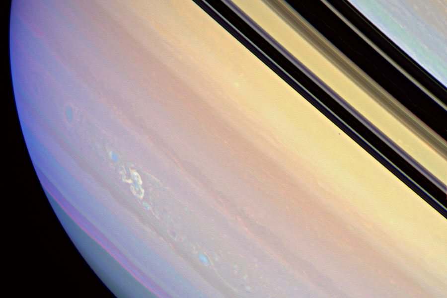 Saturn ist im Bild als Scheibe zu sehen, es verlaufen Streifen diagonal von links oben nach rechts unten, sie sind von violett bis orange-gelb schattiert, rechts oben sind die Ringe als schwarze Streifen zu sehen.