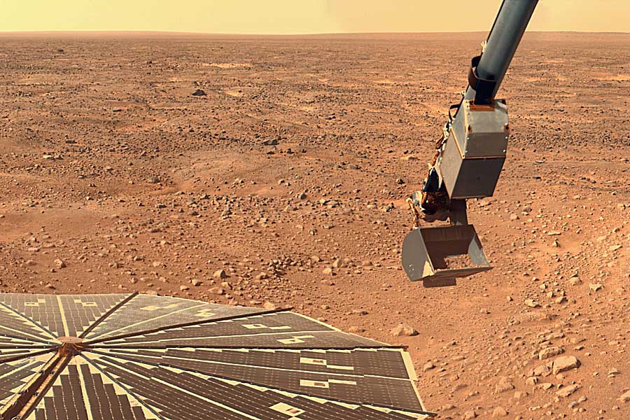 Auf dem roten, von Geröll bedeckten Marsboden befindet sich liinks eine Anordnung von Solarpaneelen, von oben ragt ein Schaufelarm ins Bild. Über dem Horizont ist der Himmel hellorange.