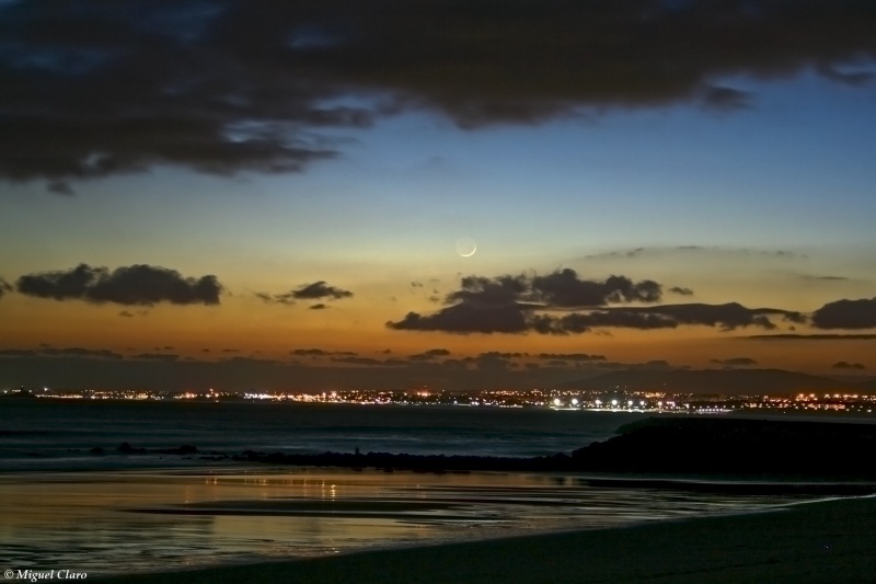 Am Abendhimmel über Lissabon leuchtet ein junger Sichelmond hinter dunklen Wolken.
