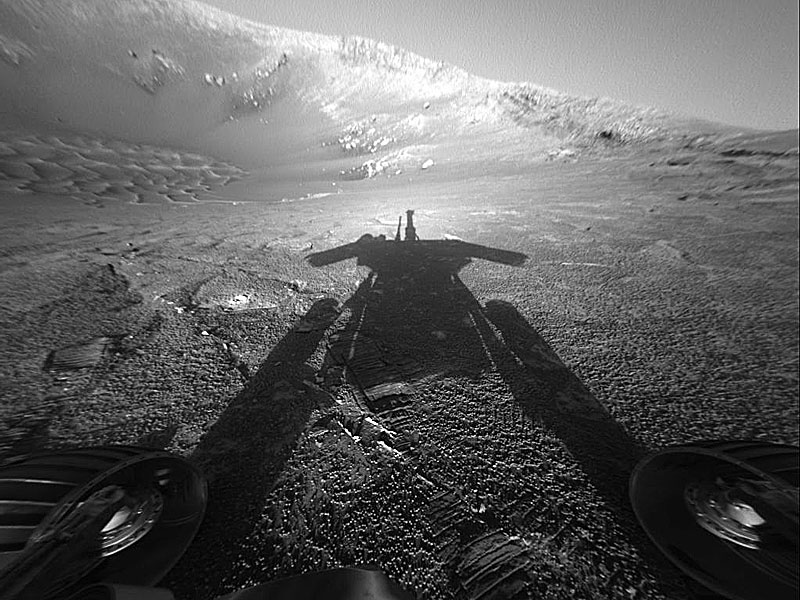 Das Schwarzweiß-Bild zeigt eine Marslandschaft, im Hintergrund ist ein Kraterrand, ins Bild fällt der Schatten des Marsrovers Opportunity.