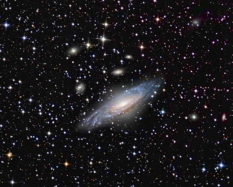 Von Sternen umgeben leuchtet eine geneigte Spiralgalaxie, darüber sind weitere, kleinere Galaxien zu sehen.