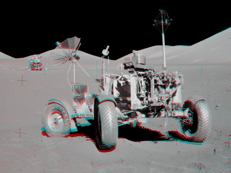 Das Bild wirkt schwarzweiß, hat aber rote und cyanfarbene Ränder. In einer Landschaft mit rundlichen Mondbergen im Hintergrund steht das Mondfahrzeug, rechts hinten ist das Apollo-Landemodul zu sehen.