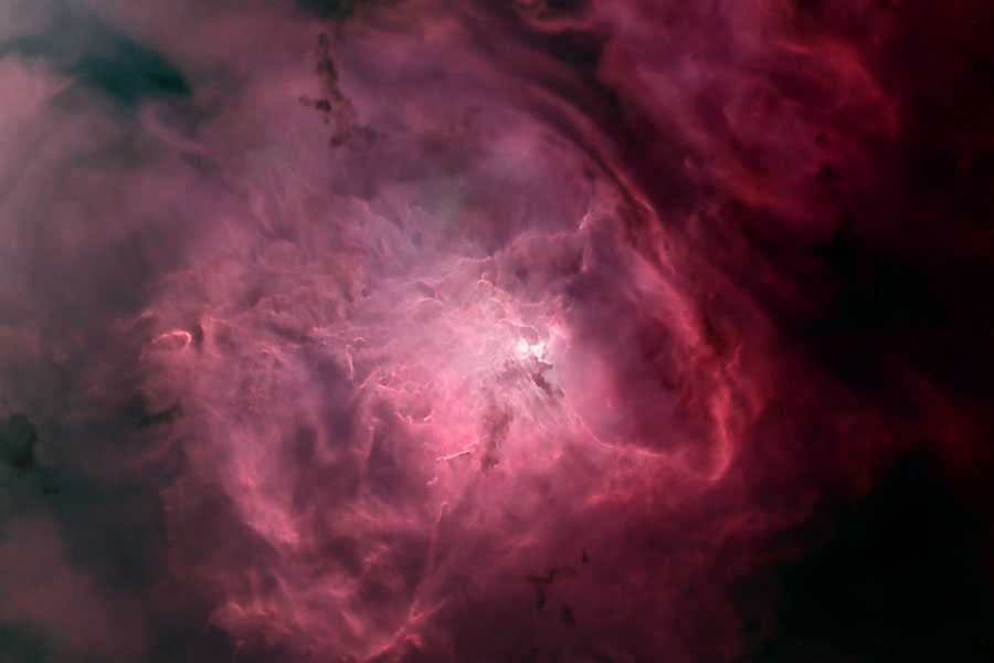 Vor einem dunklen Hintergrund leuchtet eine rötliche Gaswolke mit einigen dunklen Staubnebeln und einem hellen Zentrum.