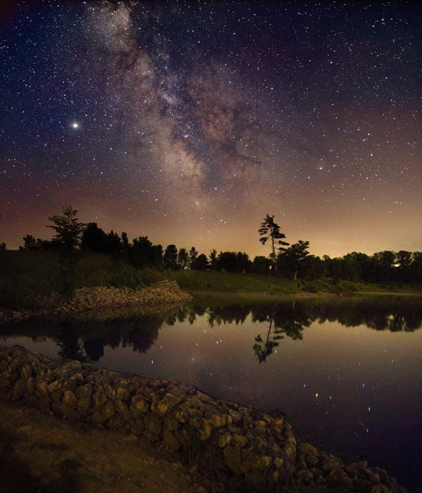 Im ruhigen See im Vordrgrund spiegelt sich der Sternenhimmel mit Milchstraße, am Ufer hinten stehen die Silhouetten von Bäumen.