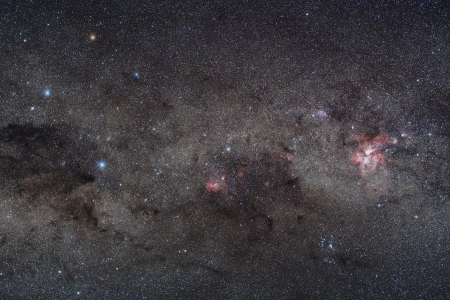 Das Bild zeigt einen Ausschnitt der Milchstraße mit Dunkelwolken, rötlichen Nebeln und blauen Sternen.