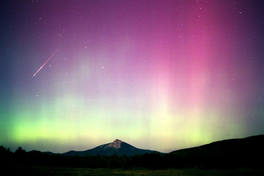 Hinter der Silhouette eines Gebirges leuchtet am Himmel ein unten grünes und oben violettes Polarlicht. Links blitzt ein Meteor.