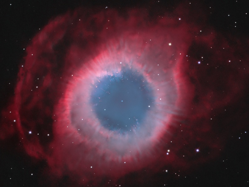 Der runde, rote Nebel hat in der Mitte ein Loch, das schwach blau leuchtet.