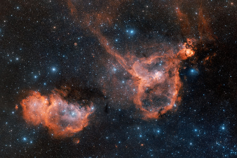 In einem Bildfeld mit blau leuchtenden Sternen leuchten zwei Nebel mit rotem Rand.