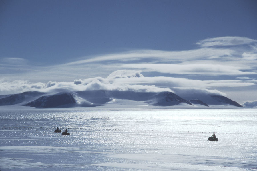 Hinter einer glänzenden Eisfläche mit einigen dunklenObjekten ist ein Gebirge zu sehen, darüber ein heiterer blauer Himmel mit einigen Wolken.