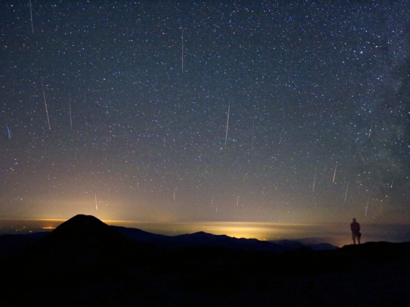 Hinter der Silhouette einer Berglandschaft mit der Silhouette eines Beobachters in der Ferne sind von unten beleuchtete Wolken am Horizont, darüber ist ein sternklarer Himmel mit zahlreichen Meteorspuren.