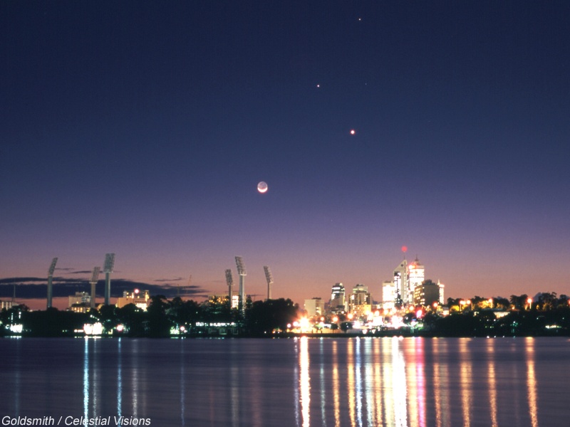 Die Silhouette von Perth spiegelt sich im Wasser im Vordergrund, darüber leuchten am dunkelblauen Himmel der Mond und die Planeten Merkur, Venus und Mars.