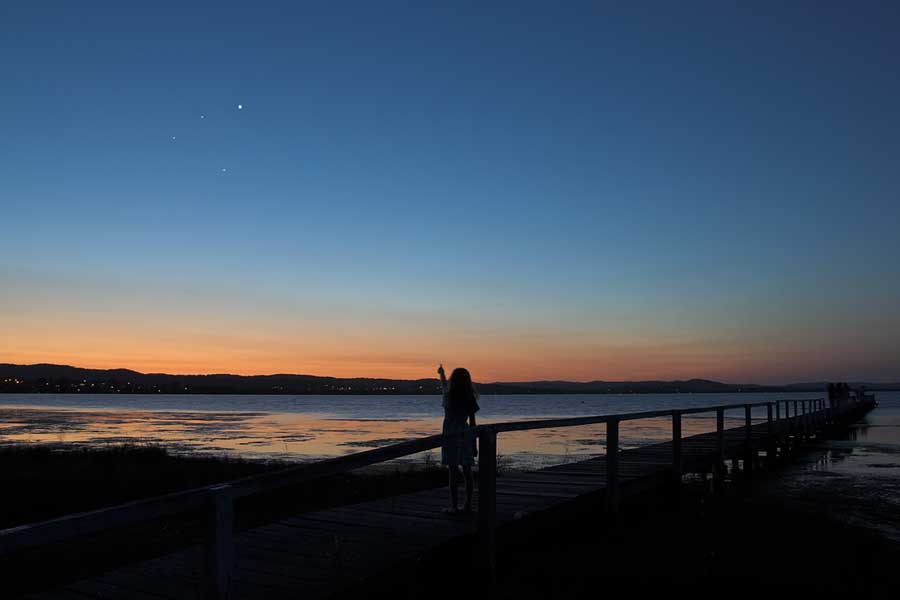 Hinter einem Gewässer leuchten am blauen Himmel drei Lichtpunkte. Der Horizont ist orangerot gefärbt, im Vordergrund sind die Silhouetten eines Ufers, eines Geländers und einer Person zu sehen.