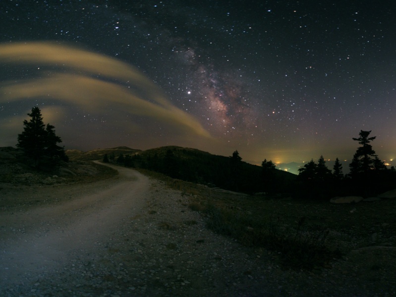 Hinter dem dunklen, silhouettenartigen Horizont leuchtet die Sommermilchstraße, Planet Jupiter, Stadtlichter im Hintergrund und links schwach beleuchtete Wolken.