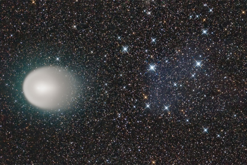 Rechts ist vor einem Sternenfeld mit einigen helleren gezackten Sternen eine weiß-graue ovale Hülle mit einem hellen Kern zu sehen. Rechts ist die Hülle leicht ausgefranst.