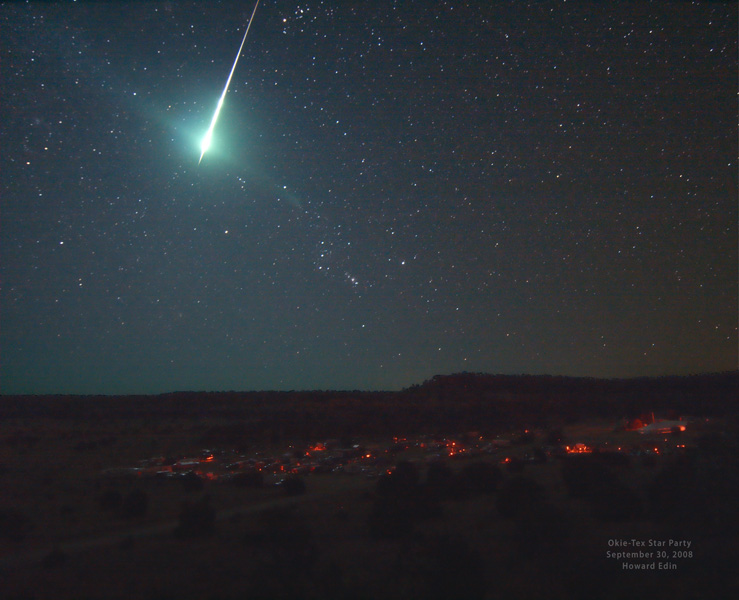 Am sternklaren Himmel blitzte eine Feuerkugel auf, am Sternenhimmel ist das Sternbild Orion zu sehen, unten ist die Silhouette einer Landschaft mit wenigen roten Lichtern.