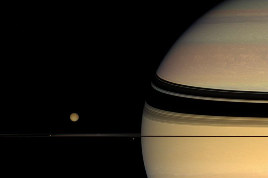 Rechts ist ein großer Teil des Planeten Saturn zu sehen, auf der orangefarbenen Oberfläche zeichnen sich die Schatten der Ringe ab, links daneben befindet sich der Saturnmond Titan. Die Ringe laufen als schmaler schwarzer Strich durchs Bild.