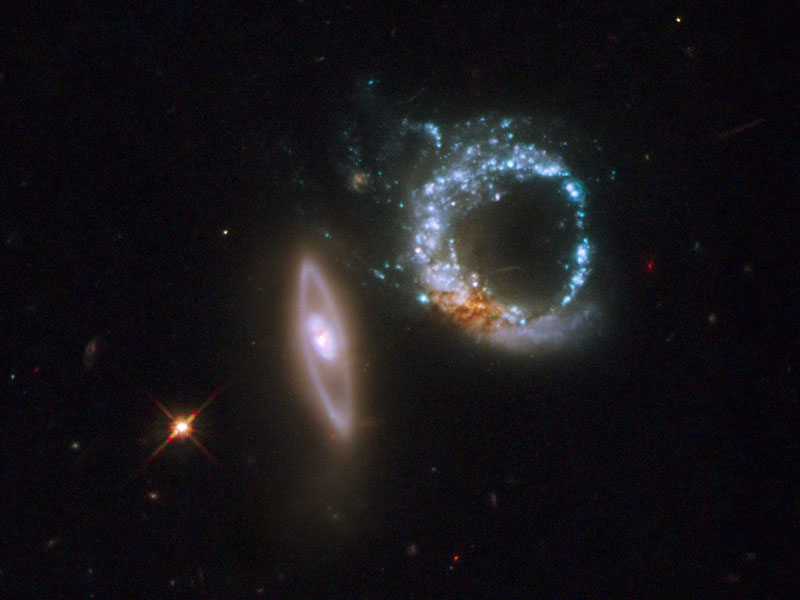 Auf einer Diagonale vor schwarzem Hintergrund ist links unten ein orangefarbener Stern, in der Mitte eine senkrechte verzerrte Galaxie und rechts oben ein blauer Ring aus Sternhaufen.