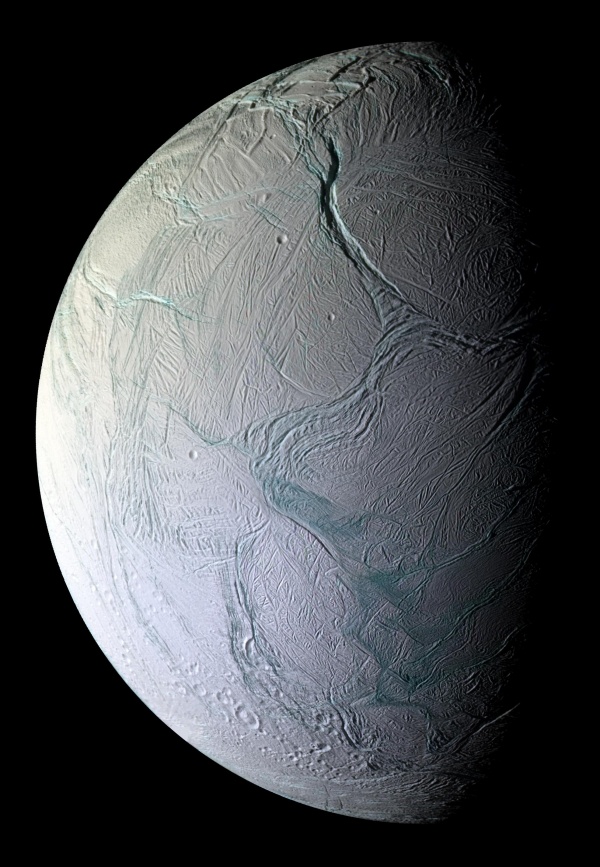 Der Mond Enceladus ist von der linken Seite zur Hälfte beleuchtet. Seine eisige Oberfläche ist von Gräben und Falten überzogen, die zum Teil einen bläulichen Farbton haben.