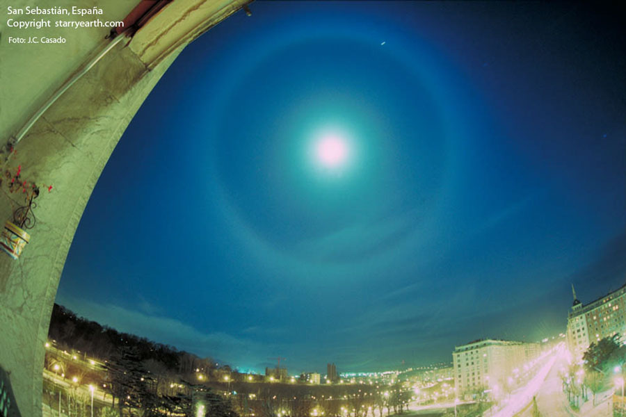 Über einer Stadt leuchtet am blauen Nachthimmel der Mond, umgeben von einem leuchtenden Halo. Auf der Ein-Uhr-Position leuchtet der Planet Jupiter.